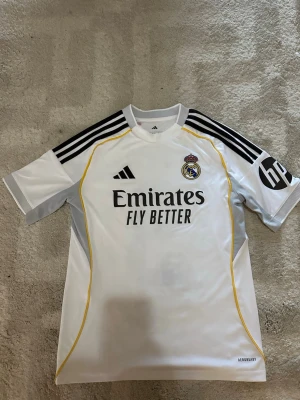 Real Madrid Mbappé matchtröja - Vit Real Madrid fotbollströja från Adidas med svarta detaljer och gula linjer. Tryck med 'Emirates Fly Better' på framsidan och 'Mbappé 9' på ryggen. Klubblogga på bröstet och svart HP-logga på ärmen. Tillverkad i lätt funktionsmaterial.