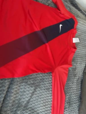 Röd långärmad Nike Dri-Fit tröja - Röd långärmad sporttröja från Nike med svart diagonal rand och vit Nike-logga på bröstet. Tillverkad i Dri-Fit material som andas och passar perfekt för träning eller fotboll. Tröjan har rund hals och är lätt och smidig.