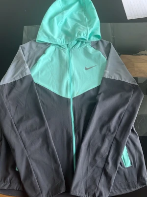 Nike vindjacka i mint och grå - Snygg vindjacka från Nike i mintgrönt och olika nyanser av grått. Jackan har huva, hel dragkedja och två sidofickor. Perfekt för blåsiga dagar och sportiga outfits. Nike-logga på bröstet och modern, sportig design.