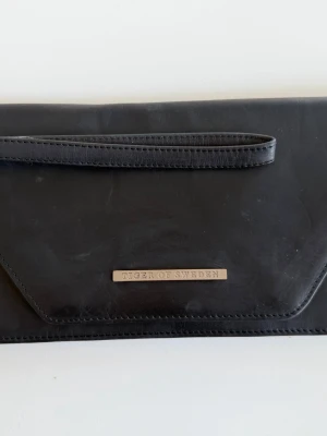 Elegant Tiger of Sweden Clutch i Äkta Läder - Stilren och tidlös svart kuvertväska från exklusiva Tiger of Sweden. Den minimalistiska designen med den eleganta V-formade fliken och diskreta metalldetaljen framtill gör väskan perfekt både till fest, middag och kvällsevent.  Väskan är tillverkad i läder och har en praktisk avtagbar handledsrem som gör den både snygg och smidig att bära. Perfekt storlek för mobil, kort, nycklar och andra små essentials.  Stilren skandinavisk design Avtagbar handledsrem Elegant metalldetalj med logotyp 