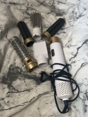 Multistyler hårverktyg med flera tillbehör - Säljer en multistyler för hår med flera utbytbara tillbehör. Inkluderar rundborste, volymborste, två locktillbehör och en fönmunstycke. Verktyget är vitt med guld- och svarta detaljer och har sladd. Perfekt för styling och olika frisyrer.