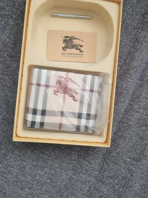 Rutig plånbok från Burberry - Snygg plånbok från Burberry i klassiskt beige, svart och rött rutmönster. Plånboken har Burberrys ikoniska ryttare tryckt på framsidan och är tillverkad i tåligt läder. Perfekt för dig som vill ha en stilren accessoar med lyxig känsla.