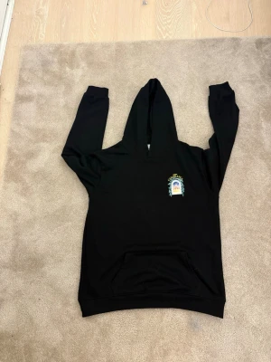 Svart hoodie från Casablanca - Svart hoodie från Casablanca med färgglatt tryck på bröstet och stor grafisk print på ryggen med texten 'Avenida Casablanca' och ett motiv av en byggnad. Klassisk känguruficka framtill och huva. Perfekt för en chill och stilren look.