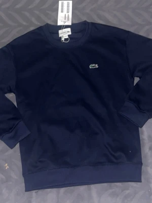 Mörkblå sweatshirt från Lacoste - Mörkblå sweatshirt från Lacoste med klassisk rund halsringning och det ikoniska gröna krokodilmärket broderat på bröstet. Tröjan har ribbade muddar vid ärmslut och nederkant. Perfekt för en stilren och avslappnad look.