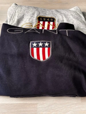 GANT hoodie med broderad logga - Två snygga hoodies från GANT, en i svart och en i ljusgrått. Båda har broderad GANT-logga och sköld med stjärnor och ränder på bröstet. Klassisk passform och mjukt material, perfekt för en avslappnad stil. Man får båda för 300 kr