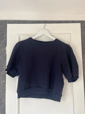 Marinblå topp från Gina Tricot - Säljer en marinblå topp från Gina Tricot med korta puffärmar och ribbade muddar. Toppen är croppad och gjord i mjuk bomull, perfekt för en avslappnad och trendig look. I storlek 146/152