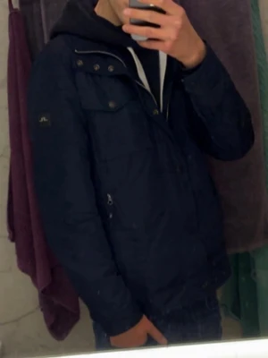 J Lindberg field jacket  - Stilren mörkblå vindjacka från j Lindberg  med hög krage, dragkedja och tryckknappar. Jackan har två bröstfickor med lock och två sidofickor med dragkedja. Perfekt för blåsiga dagar och enkel att matcha med olika outfits.