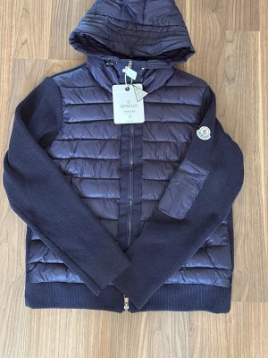 Moncler Cardigan - Snygg mörkblå pufferjacka från Moncler med quiltad framsida och huva. Jackan har stickade ärmar och sidor, dragkedja framtill och en Moncler-logga på ärmen. Perfekt för kyliga dagar och riktigt trendig look.