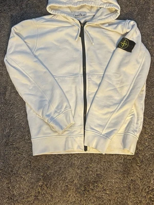 Vit hoodie från Stone Island - Säljer en vit hoodie från Stone Island med dragkedja framtill och klassisk huva med dragsko. På vänster ärm sitter det ikoniska svarta Stone Island-märket med gul och grön brodyr. Hoodien har ribbade muddar och är tillverkad i mjukt bomullsmaterial.