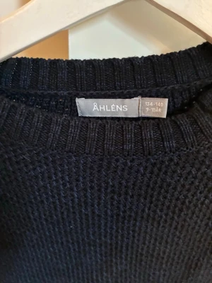 Mörkblå stickad tröja från Åhléns - Mörkblå stickad tröja från Åhléns med rund halsringning och ribbade muddar vid ärmslut och nederkant. Tröjan har en klassisk, enkel design och är perfekt för att hålla värmen under kyliga dagar.