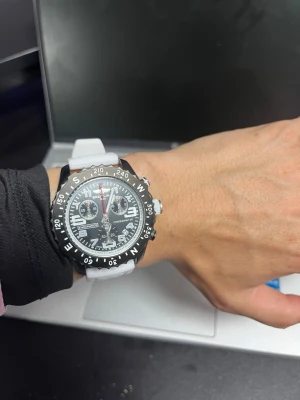 Svart och vit Breitling klocka - Snygg Breitling klocka med svart urtavla, vita detaljer och vit silikonrem. Klockan har flera funktioner som kronograf och datumvisning samt tydliga markeringar på boetten. Perfekt accessoar för dig som gillar sportig och modern stil.