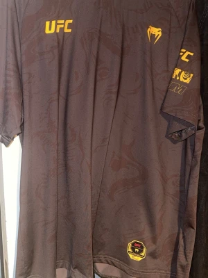 Svart UFC t-shirt från Venum - Säljer en svart t-shirt från Venum med UFC-logga i gult på bröstet och ärmen. Tröjan har diskret mönster i tyget och är tillverkad i ett lätt och snabbtorkande material, perfekt för träning eller sport.