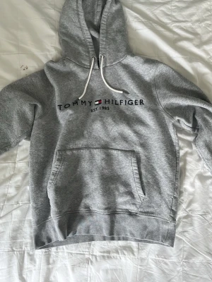 Grå hoodie från Tommy Hilfiger - Snygg grå hoodie från Tommy Hilfiger med broderad logga och flagga på bröstet. Klassisk känguruficka framtill, vit dragsko i huvan och mjukt fleecefoder på insidan. Perfekt för en chill och stilren look.
