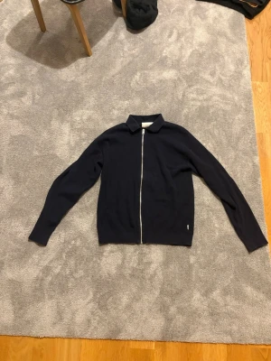 Mörkblå ziptröja med krage - Snygg mörkblå ziptröja med klassisk krage och hel dragkedja framtill. Tröjan har lång ärm och är ribbstickad för en stilren look. Perfekt att slänga på sig över en t-shirt för en avslappnad men trendig stil.