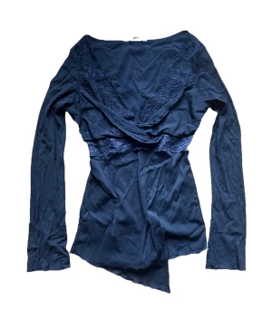 Y2k vintage Svart omlottopp med spetsdetaljer - Trendig svart omlottopp med djup V-ringning och fina spetsdetaljer längs halsringningen och under bysten. Toppen har långa ärmar och asymmetrisk nederkant som ger en cool look. Perfekt att styla med jeans eller kjol.