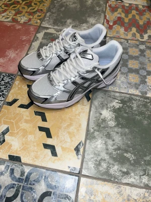 Asics Gel-1130 Pure Silver - Snygga Asics sneakers i vitt och silver med svarta detaljer. Skorna har mesh- och syntetmaterial för en sportig look och bra andningsförmåga. Klassisk låg modell med snörning och bekväm sula, perfekta för en trendig streetstyle.