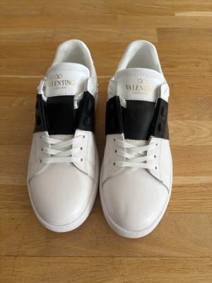 Valentino skor  - Snygga vita sneakers från Valentino Garavani med svart detalj över snörningen och guldfärgad logga på plösen. Skorna är låga och har en klassisk siluett i slätt läder med vit sula och diskret mönstrad undersida. Perfekta för dig som gillar stilrena och exklusiva sneakers.