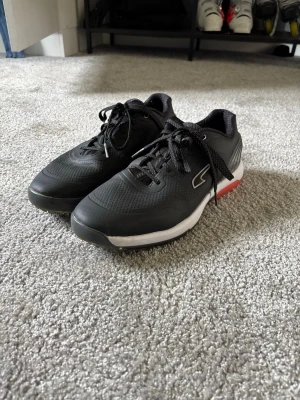 Svarta Puma golfskor med röd detalj - Svarta sneaker-golfskor från Puma med vit och röd sula. Skorna har en låg profil, snörning och meshdetaljer för bra ventilation. Snygg sportig look med puma-logga på sidan. Materialet är en mix av syntet och mesh som ger en lätt känsla. Helt nya.