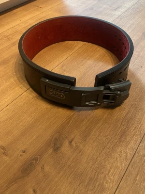 SBD svart träningsbälte i skinn - Säljer ett svart träningsbälte från SBD med röd insida. Bältet är tillverkat i kraftigt skinn och har ett robust spänne i metall. Perfekt för styrketräning och gym. Klassisk design med justerbara hål för optimal passform. Pris kan diskuteras!!