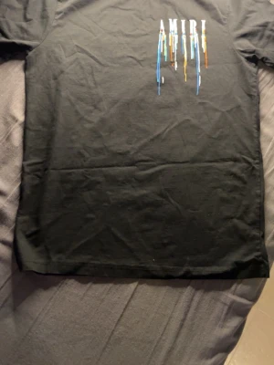 Svart AMIRI t-shirt med färgstänk - Säljer en svart t-shirt från AMIRI med coolt tryck framtill där loggan har färgstänk i blått, vitt, orange och gult som rinner ner. Klassisk passform och rund hals. Perfekt för dig som vill ha en unik och trendig look.