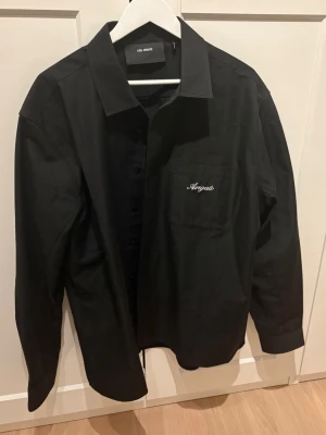 Svart overshirt från Axel Arigato - Svart overshirt från Axel Arigato med klassisk krage, knappstängning och bröstficka med vit broderad logga. Overshirten har en clean och stilren look, perfekt att bära öppen eller stängd. Knappt använd, nypris över 2500:-