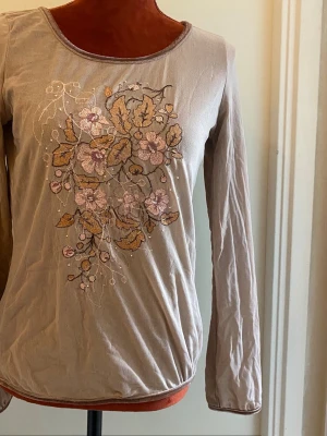 Beige långärmad topp med broderi - Beige långärmad topp med rund halsringning och broderade blommor i rosa och gult på framsidan. Tunn och mjuk känsla, perfekt för dig som gillar romantiska detaljer och en feminin look.