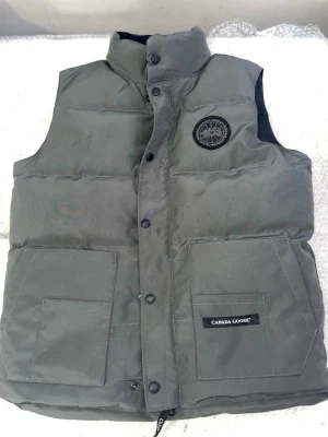 Grå dunväst från Canada Goose - Snygg grå dunväst från Canada Goose med hög krage och två stora framfickor. Västen har ett klassiskt märke på bröstet och stängs med tryckknappar. Tillverkad i slitstarkt material och fylld med dun för extra värme.