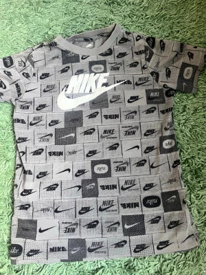 Grå Nike t-shirt med logotypmönster - Säljer en grå t-shirt från Nike med korta ärmar och all-over print av olika Nike-logotyper i svart och vitt. T-shirten har en klassisk passform och är perfekt för dig som gillar sportiga och trendiga plagg. Materialet känns mjukt och bekvämt. Hör av er vid minsta lilla fråga. Priset är inte hugget i sten.