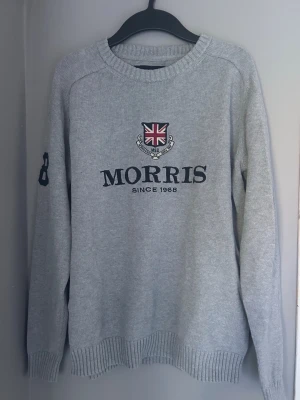 Grå stickad tröja från Morris - Snygg grå stickad tröja från Morris med broderad Union Jack och text på bröstet. Tröjan har ribbade muddar, rund hals och siffran 68 på ena ärmen. Perfekt för dig som gillar klassisk och sportig stil. Perfekt för de sena sommarkvällarna som är nära!☀️