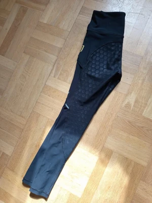 Svarta sportleggings från Maya delorez  - Säljer ett par svarta sportleggings från Maya delorez med hög midja och diskreta mönsterdetaljer längs benen. De har gråa sömmar och är tillverkade i ett stretchigt funktionsmaterial som passar perfekt för träning.