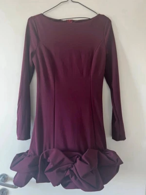 Vinröd klänning med volangkant - Säljer en vinröd klänning med långa ärmar och rund halsringning. Klänningen har en unik volangkant nedtill som ger en cool och trendig look. Materialet är stretchigt och följsamt, perfekt för dig som vill sticka ut lite extra. Oanvänd 