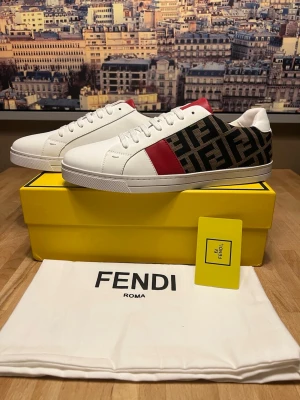 Fendi vita och svarta sneakers - Snygga sneakers från Fendi i vitt läder med svart textilpanel med FF-logga och en röd detalj på sidan. Klassisk låg modell med vita snören och gummisula. Perfekta för dig som vill ha en exklusiv och trendig look.