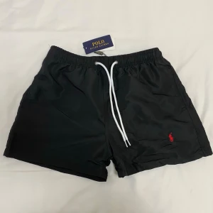 Svarta badbyxor från Polo Ralph Lauren - Svarta badbyxor från Polo Ralph Lauren med vit dragsko i midjan och röd broderad logga på ena benet. Klassisk modell med elastisk midja, sidfickor och en bakficka. Perfekta för stranden eller poolen.