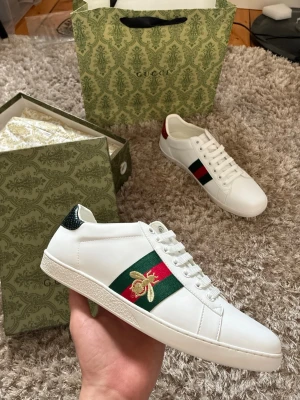Gucci Ace vita sneakers med bi - Gucci Ace sneakers i vitt läder med klassiska gröna och röda sidoremmar och broderad guldfärgad bi. Skorna har vita snören, låg profil och detaljer i metallic på hälen. Perfekta för dig som vill ha en ikonisk och lyxig sneaker med unik design.