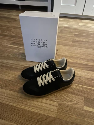 Maison Margiela Gats - Svarta sneakers från Maison Martin Margiela med vita snören och beige innersula. Klassisk låg modell med ovandel i läder och mocka, samt gummisula. Snygg och stilren design som passar till det mesta. passar 41 också