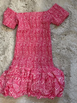 Rosa offshoulder klänning med volang - Söt rosa kortklänning med vitt bladmönster och volang nertill. Offshoulder-modell med korta ärmar, perfekt för varma dagar. Klänningen har en smickrande passform och är lätt att styla med sneakers eller sandaler.