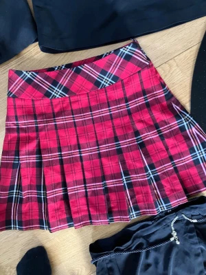 Röd och svart rutig plisserad kjol - Snygg röd och svart rutig kjol med plisserad design och klassiskt rutmönster. Kjolen har en markerad midja och är kort i modellen, perfekt för en trendig look. Passar dig som gillar färg och mönster i din stil.