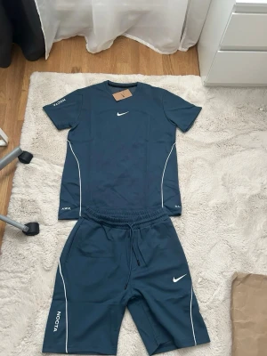 Nike NOCTA blått träningsset - Säljer ett blått träningsset från Nike NOCTA med t-shirt och shorts. Setet har vita detaljer och loggor, samt text på ärm och ben. Materialet är mjukt och stretchigt, perfekt för träning eller sportaktiviteter.