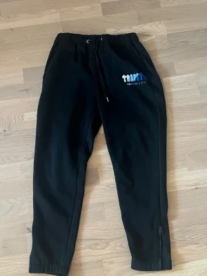 Trapstar byxor - Säljer ett par svarta mjukisbyxor från Tempstar med broderad logga i vitt och blått på ena benet. Byxorna har dragsko i midjan, fickor och dragkedja vid benslut. Perfekta för chill eller träning. Ingår hoodie