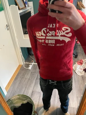 Röd Superdry hoodie med tryck - Snygg röd hoodie från Superdry med stort vitt tryck på bröstet och japanska tecken. Klassisk känguruficka framtill, ribbade muddar och dragsko i huvan. Perfekt för en avslappnad och trendig stil. Mjuk bomullsblandning som känns skön mot huden.