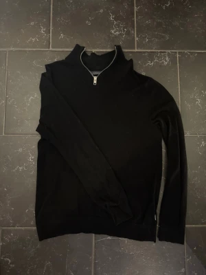 Svart halvzip tröja med hög krage - Stilren svart tröja med halvzip och hög krage. Tröjan har långa ärmar och ribbade muddar vid ärmslut och nederkant. Perfekt för lager på lager och enkel att matcha med olika outfits.