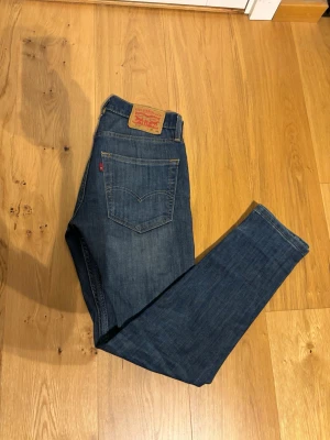 Levi's blå raka jeans - Klassiska blå jeans från Levi's med rak passform och fem fickor. Jeansen har en mörk tvätt, tydliga sömmar och den ikoniska läderlappen bak i midjan. Tillverkade i slitstarkt denim, perfekta för en avslappnad och tidlös stil.