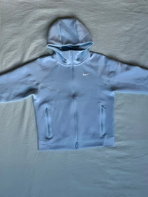 Ljusblå hoodie från Nike - Snygg ljusblå hoodie från Nike med dragkedja framtill och två fickor med dragkedja. Klassisk huva och diskret Nike-logga på bröstet. Tillverkad i mjukt material som känns bekvämt och sportigt.