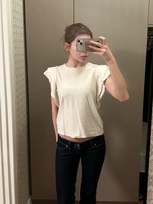 Krämvit topp med volangärm - Säljer en krämvit topp med volangdetaljer på ärmarna. Toppen har rund halsringning och en liten knappöppning i nacken. Perfekt till jeans eller kjol för en fräsch look.