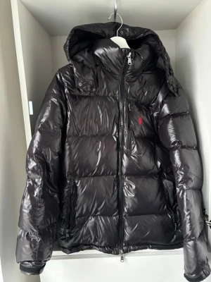 Svart pufferjacka från Polo Ralph Lauren - Säljer en svart pufferjacka från Polo Ralph Lauren med blank finish och röd logga på bröstet. Jackan har huva, dragkedja framtill och flera fickor med dragkedja. Perfekt för kalla vinterdagar och riktigt snygg att bära.