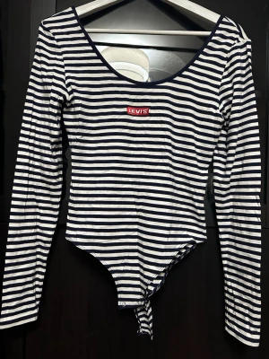 Randig långärmad body från Levi's - Säljer en snygg långärmad body från Levi's med marinblå och vita ränder. Bodyn har rund halsringning och klassisk Levi's-logga framtill. Tillverkad i mjukt bomullsmaterial som känns skönt mot huden. Perfekt för dig som gillar en enkel men stilren look.