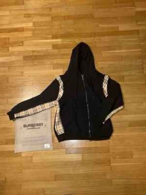 Svart Burberry hoodie med rutiga detaljer - Säljer en svart hoodie från Burberry med dragkedja och klassiska beige-rutiga detaljer längs ärmar och sidor. Hoodien har snörning i huvan och ribbade muddar. Materialet är mjukt och skönt, perfekt för dig som gillar streetwear och exklusiv design. Säljer min svarta rutiga Burberry zip eftersom den är för stor för mig. Dm mig för fler frågor.