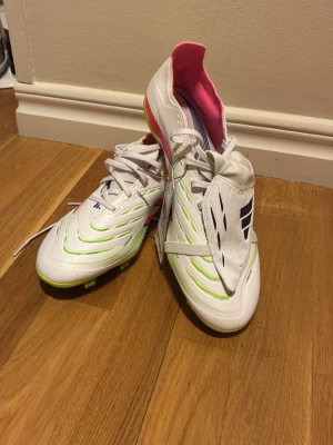 Adidas Predator vita fotbollsskor - Helt oanvända och snygga Adidas Predator fotbollsskor i vitt med neonrosa och limegröna detaljer samt mörkblå logga. Skorna har klassisk snörning, låg profil och syntetmaterial. Insidan är fodrad i rosa och yttersulan är designad för bra grepp på planen.  Oanvänd Predator League Fold-Over Tongue FG/MG, fotbollsskor gräs och konstgräs - Vit adidas   Användare: Unisex Stöd: Konstgräs, Gräs