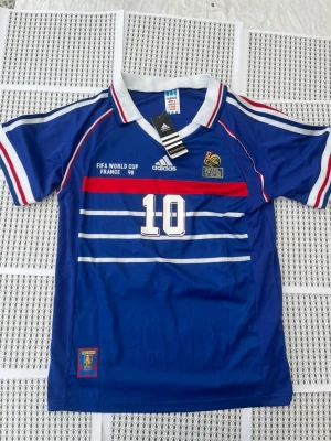 Frankrike Zidane tröja VM 98 Adidas - Blå fotbollströja från Adidas med Frankrikes landslagsmärke, nummer 10 och Zidane på ryggen. Tröjan har vita och röda detaljer, korta ärmar och klassisk krage. Materialet är lätt och andas, perfekt för sport eller samling.