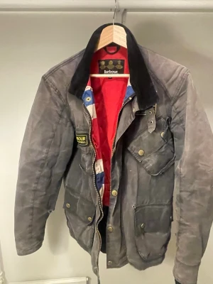 Grå Barbour jacka - Klassisk grå jacka från Barbour med svart krage i manchester, flera fickor med tryckknappar och patchdetaljer. Insidan har röd och blå färg med mönster. Perfekt för dig som gillar tidlös brittisk stil. XS passar som S